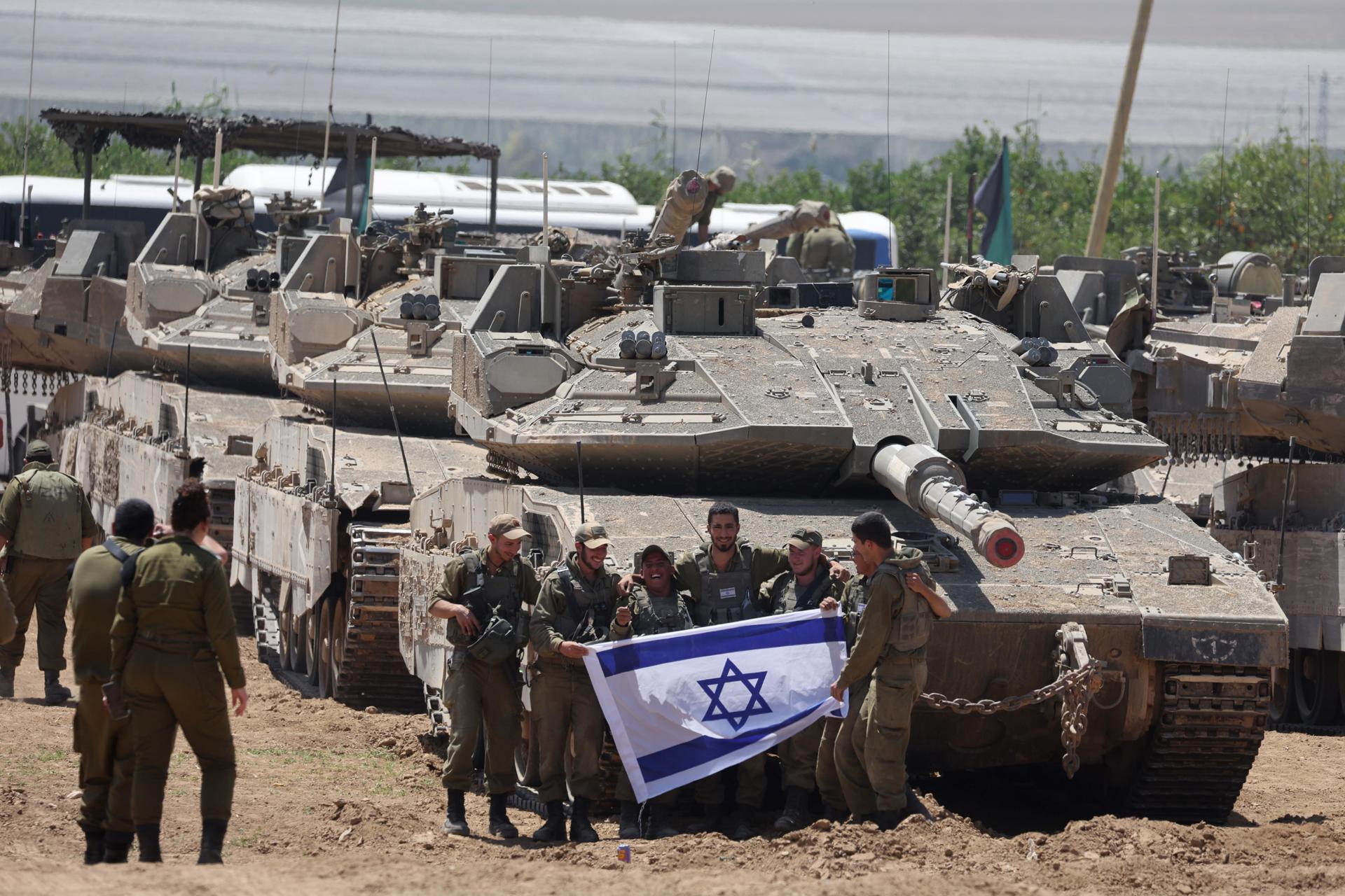 Un grupo de soldados israelíes sostiene una bandera israelí mientras se reúnen con vehículos militares en Gaza.