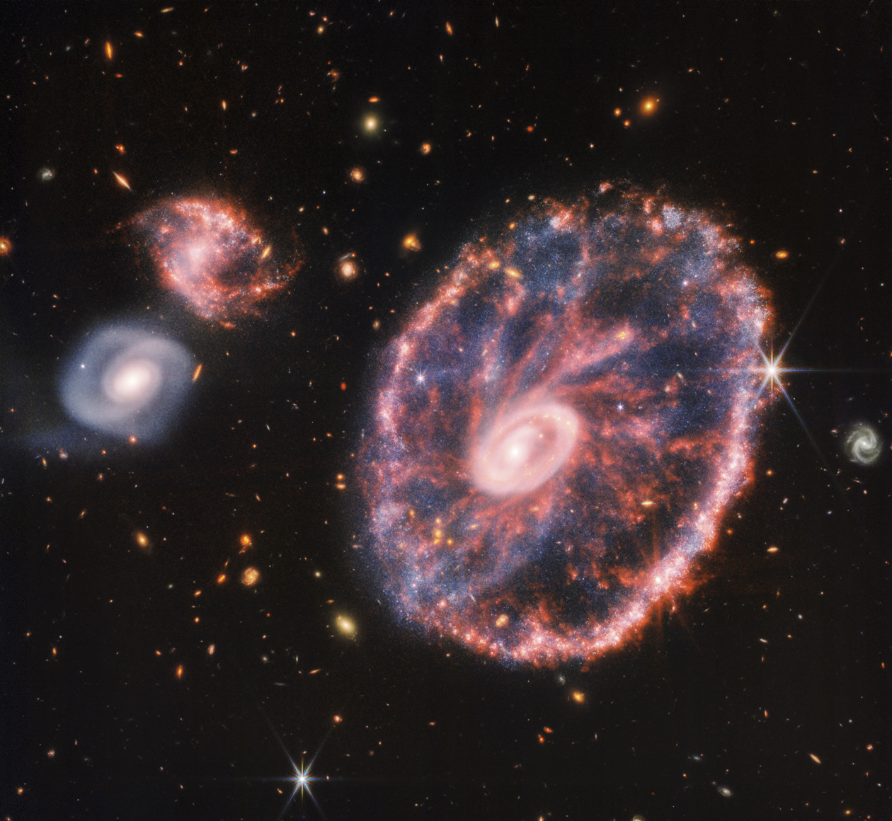 La potente mirada infrarroja del telescopio James Webb ha logrado una instantánea detallada de 'rueda de carro', pero también de dos galaxias compañeras más pequeñas con un telón de fondo de muchas otras galaxias. 