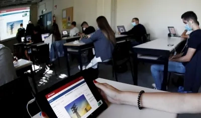 Varios alumnos siguen con ordenadores y a través de una pantalla la clase que esta impartiendo una profesora de forma presencial a otro grupo de alumnos universitarios en otro aula.